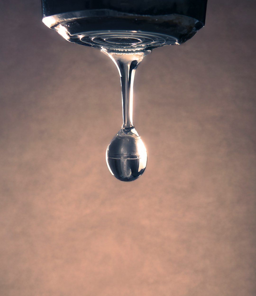 About A high-resolution macro photograph capturing a single water droplet suspended in time, hanging from the bottom of a faucet. The image is artistically flipped upside down, emphasizing the tension and fragility of the moment just before the drop falls. Warm, diffused lighting in the background adds contrast and elegance, making it ideal for use in creative projects focused on themes like time, fragility, nature, or calmness.